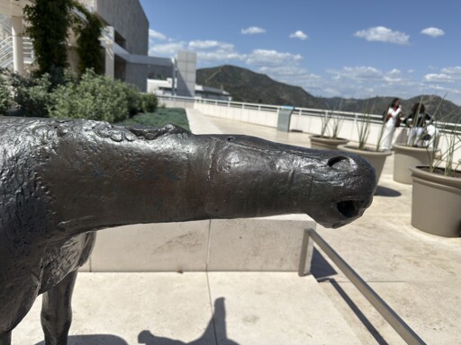 A bronze statue of the head of a horse. It is stretched out almost completely horizontally, eyes simplified to dots, and ears not visible, implying them pinned against the head. The mouth and nostrils are large, making it look as if it’s smelling something. 