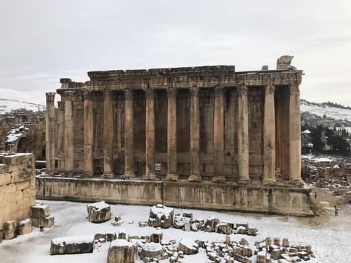 One of the long sides of the temple, roughly 20 meters tall, 60 meters long, and composed of a stone foundation supporting 15 columns, with a solid stone wall behind the columns. The surrounding landscape is littered with ruins and covered with a dusting of snow. Modern buildings are visible in the background.